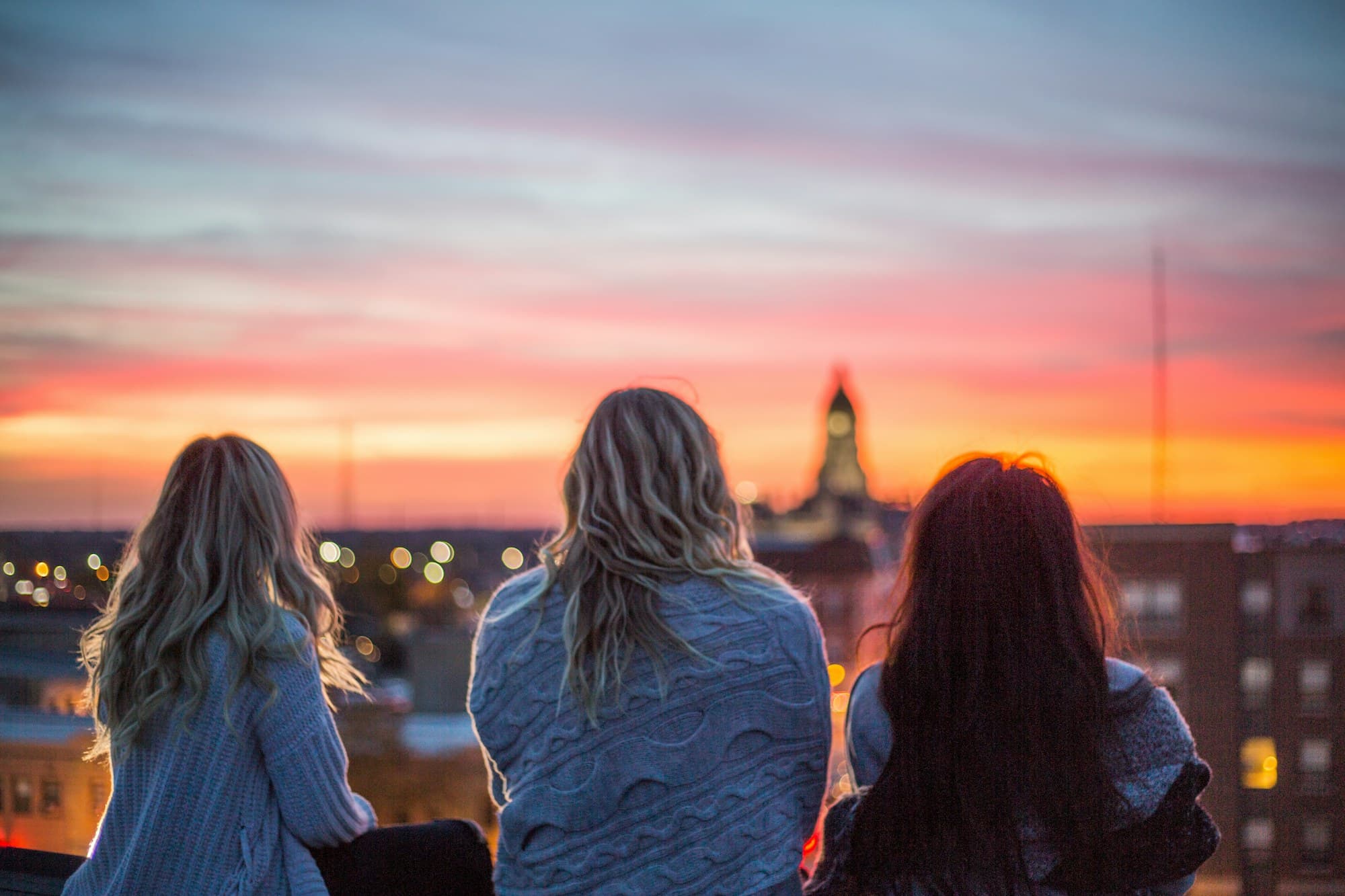 Three friends seen from behind on a rooftop, watching a pink and orange sunset over the city